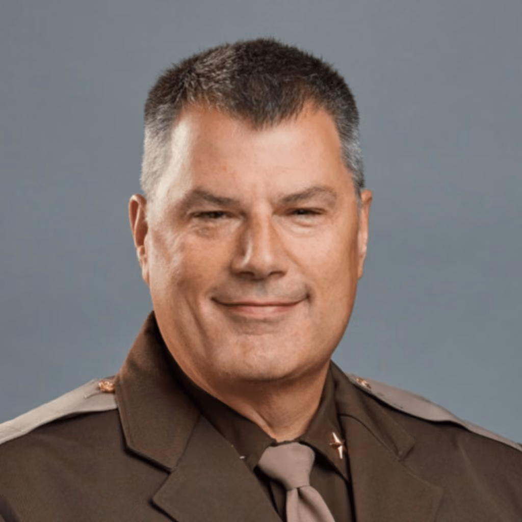 Portrait of a man in a brown police uniform with short gray hair and a slight smile.