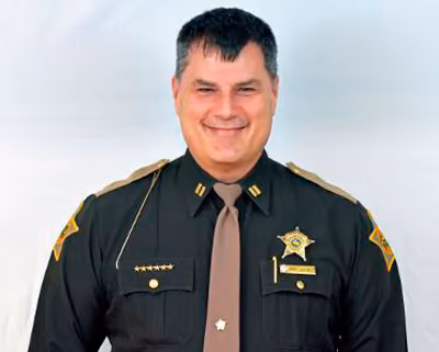 Smiling male sheriff in uniform with badges and a brown tie against light background.