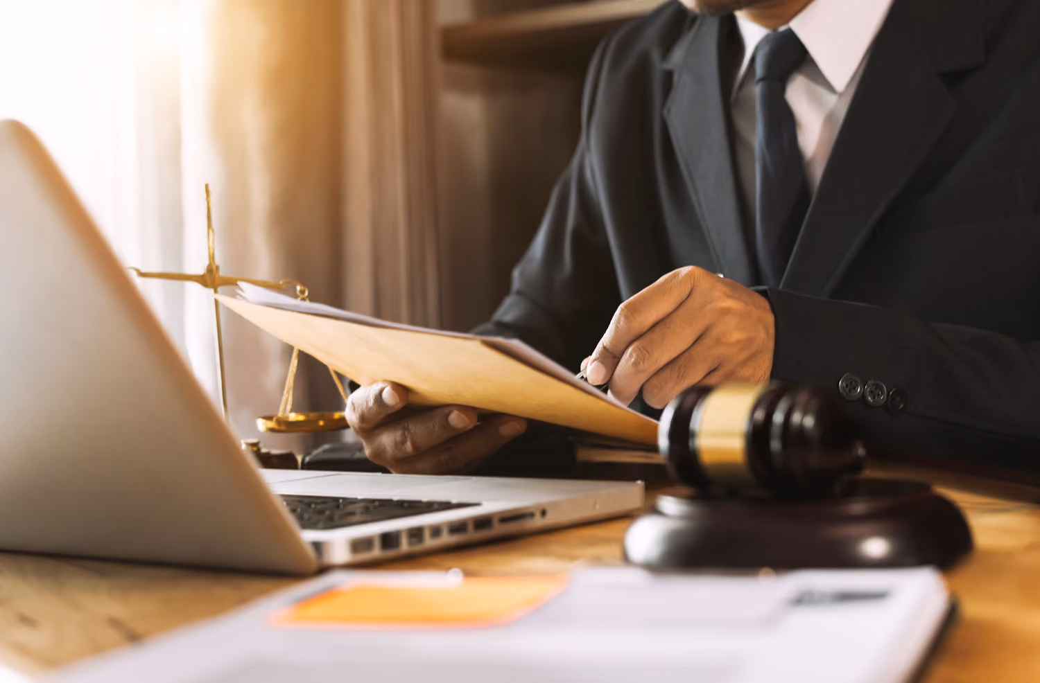 Person in suit reviewing documents at desk with laptop, gavel, and scales of justice nearby.