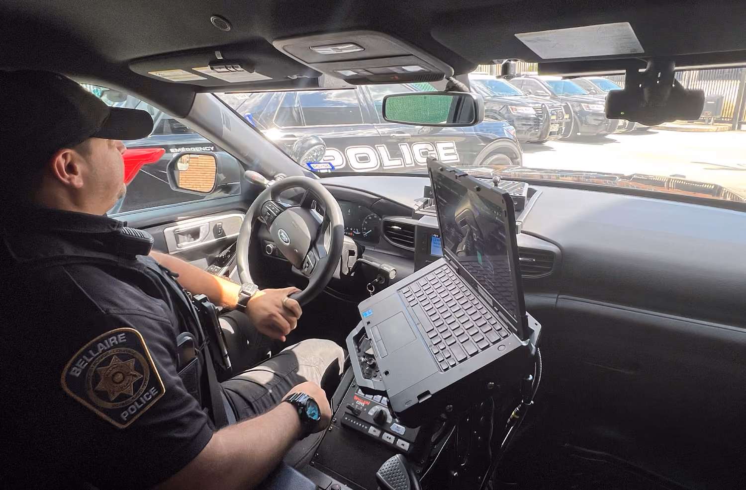 Police officer seated inside patrol car with laptop mounted on center console and other police cars outside.