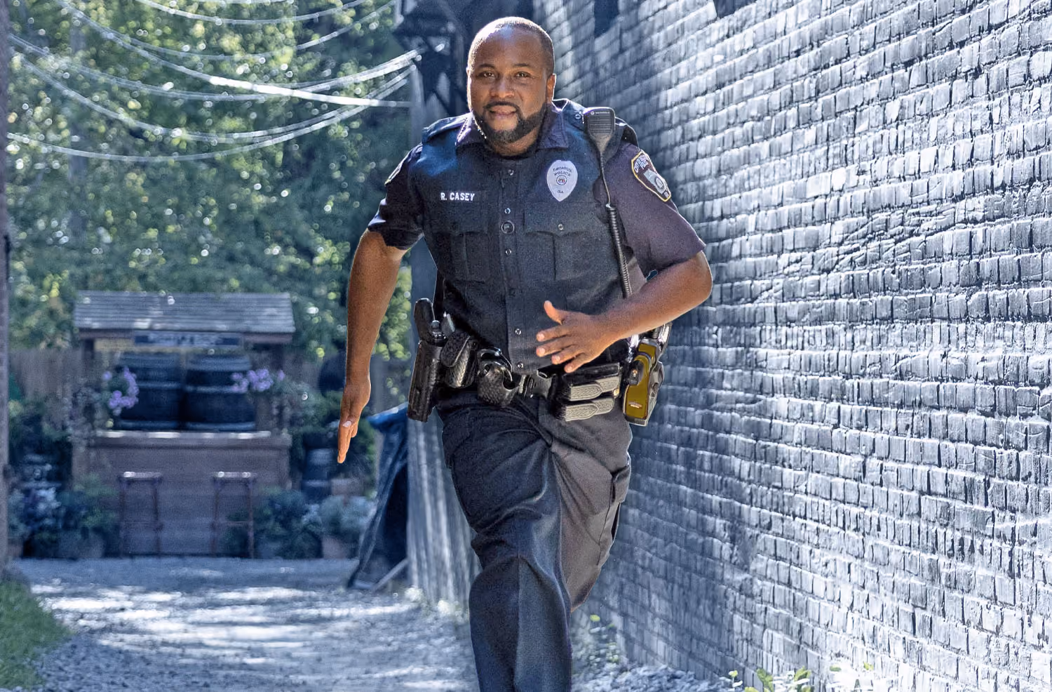 Uniformed police officer running down a narrow alley beside a brick wall during daytime.