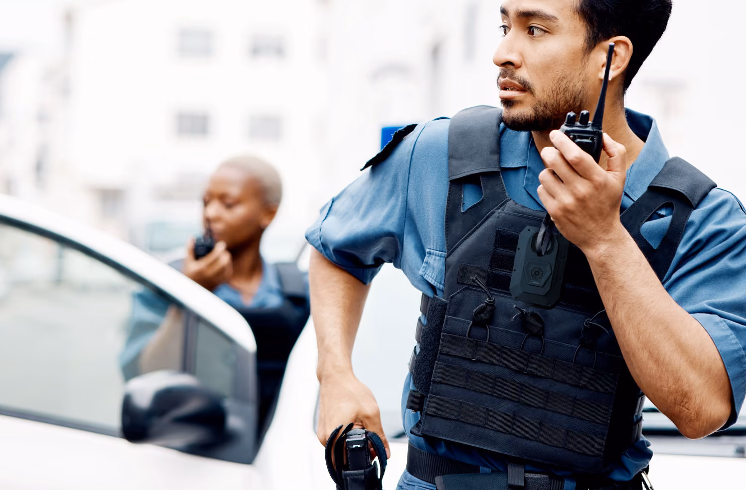 Two police officers wearing bulletproof vests using walkie-talkies near a white car outdoors.