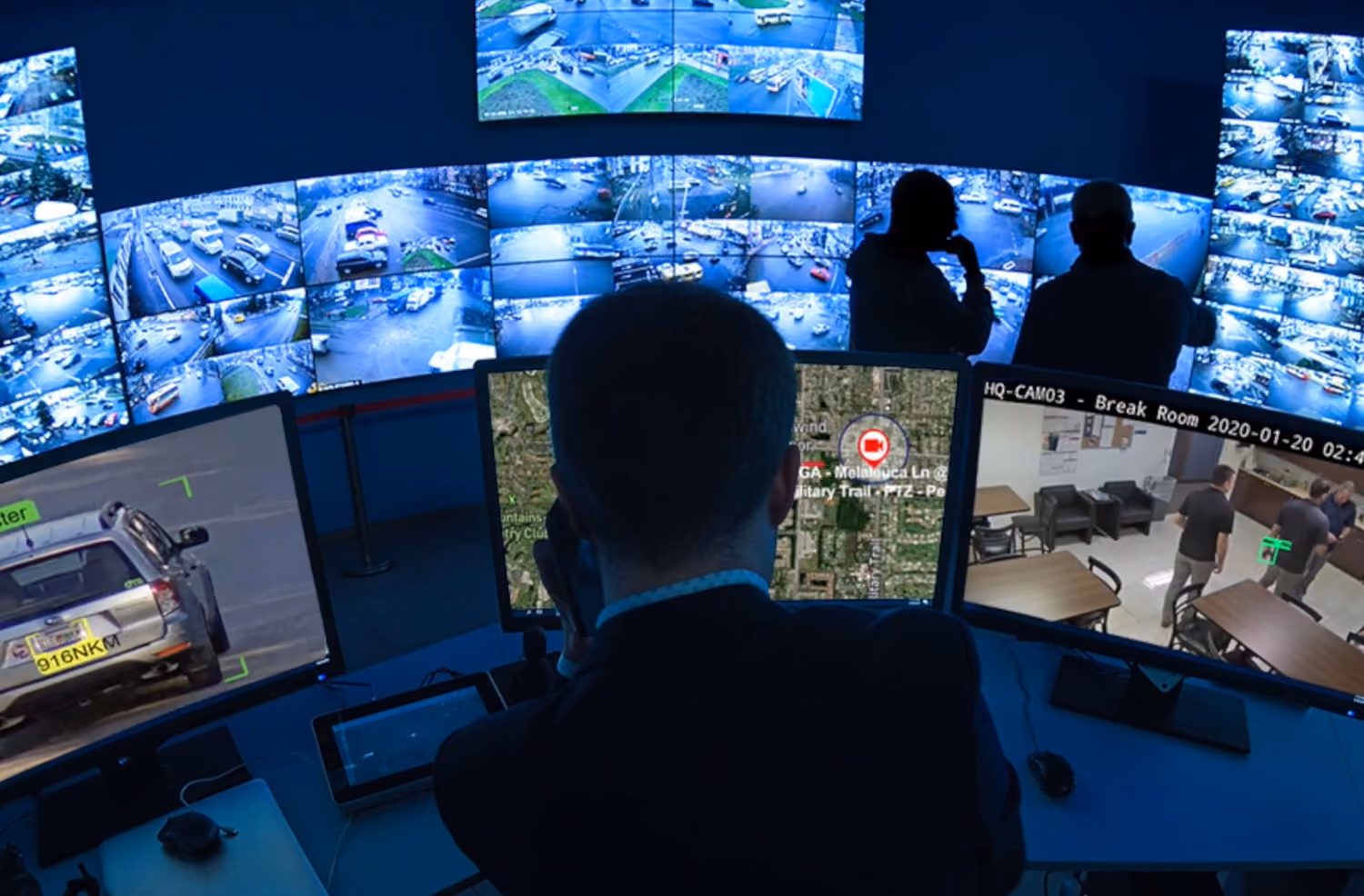 Person monitoring multiple surveillance screens showing traffic, maps, and indoor security footage.