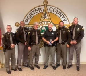 Six police officers in uniform standing in front of Porter County Sheriff's emblem on wall.