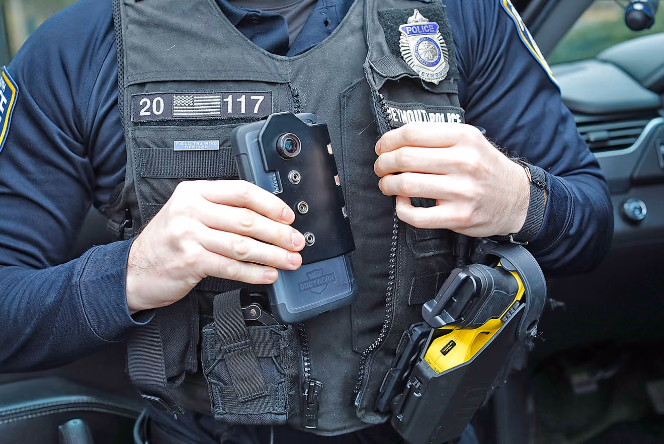 Police officer wearing tactical vest holding a body camera and a yellow Taser device, seated in a vehicle.
