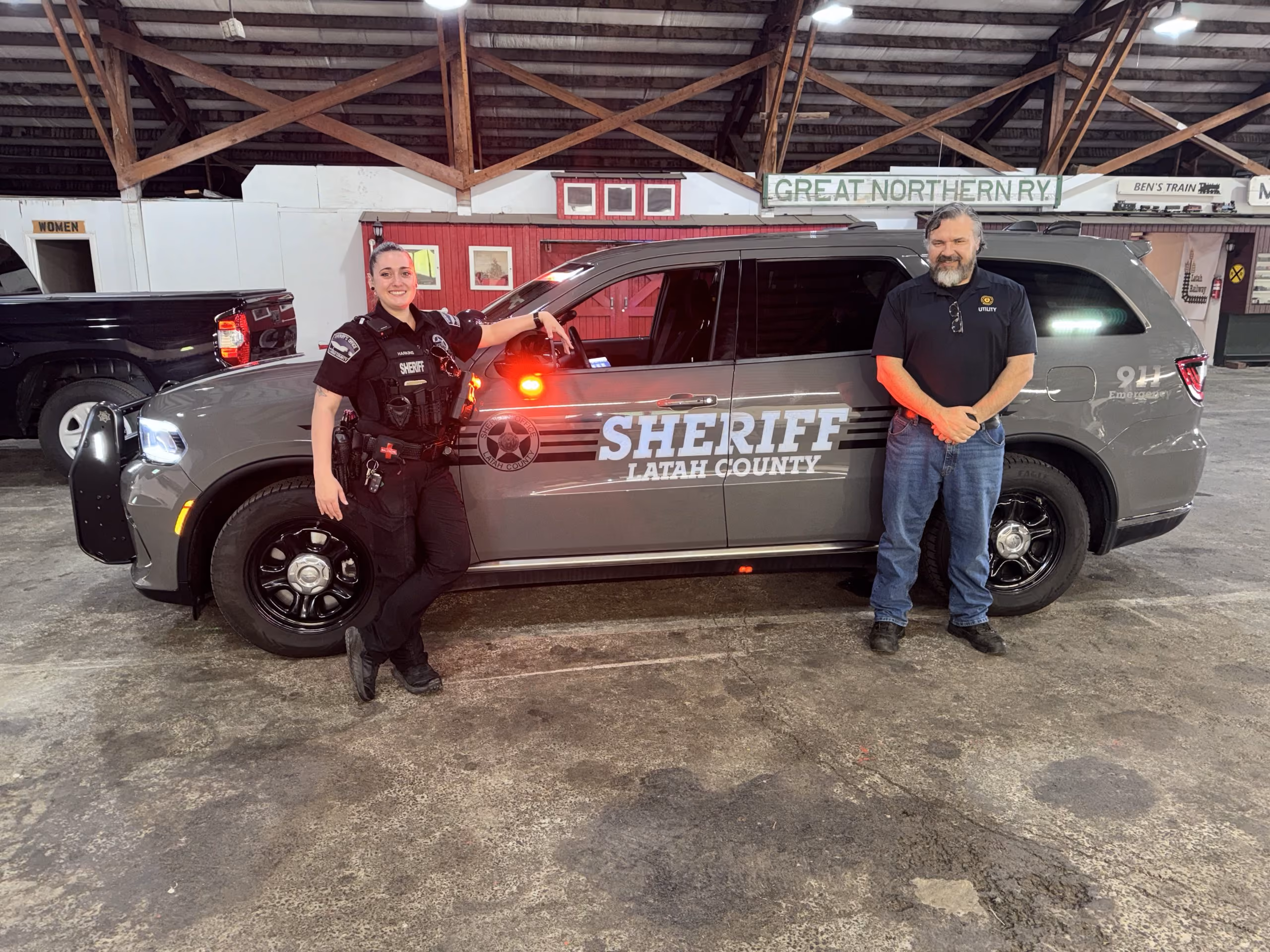 Female sheriff in uniform and man in utility shirt pose beside a Latah County sheriff SUV with red lights on.