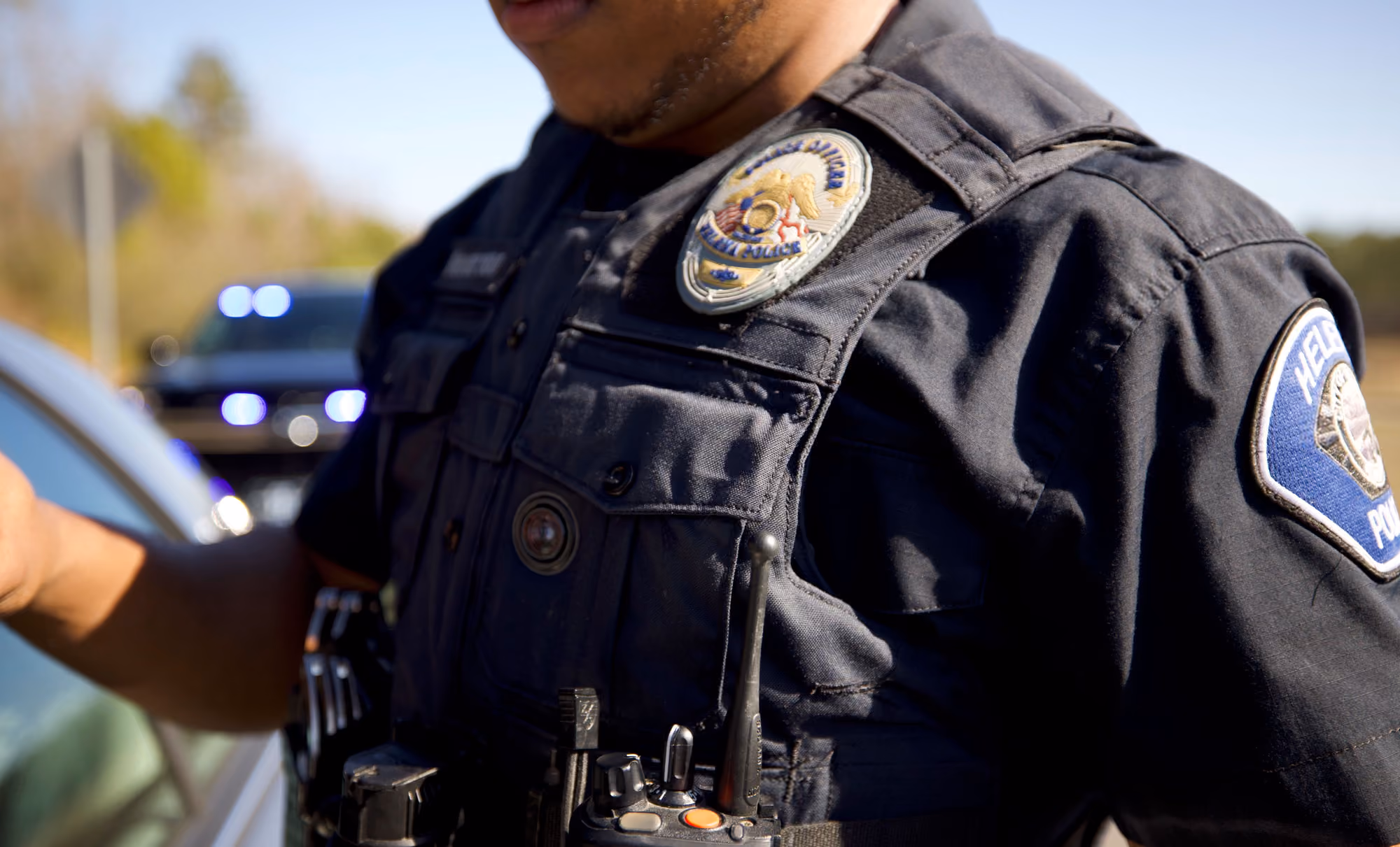 Police officer in dark uniform with badge and radio, standing near car with police lights in background.