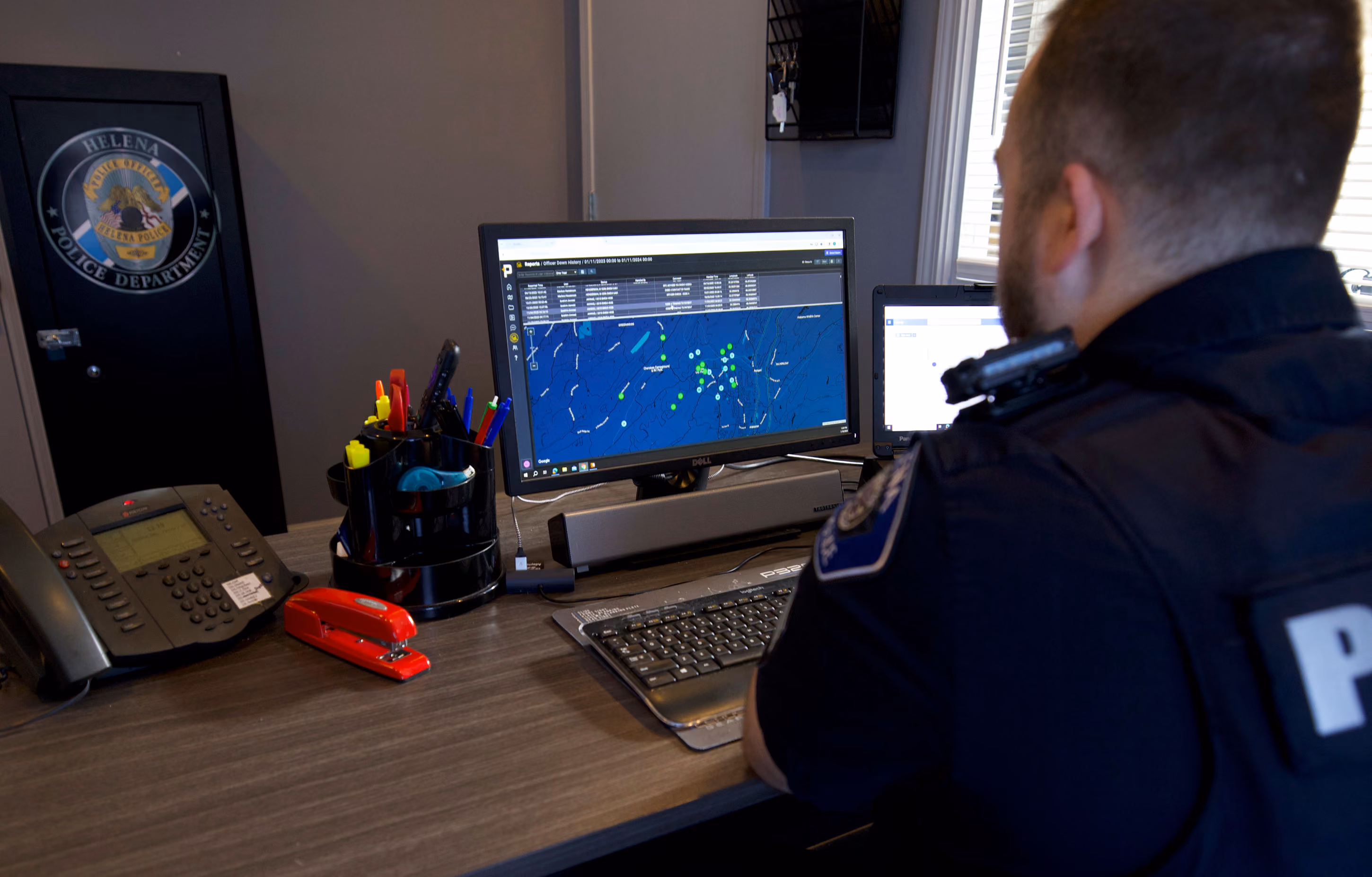 Police officer at desk working on dual monitors showing map and data in Helena Police Department office.