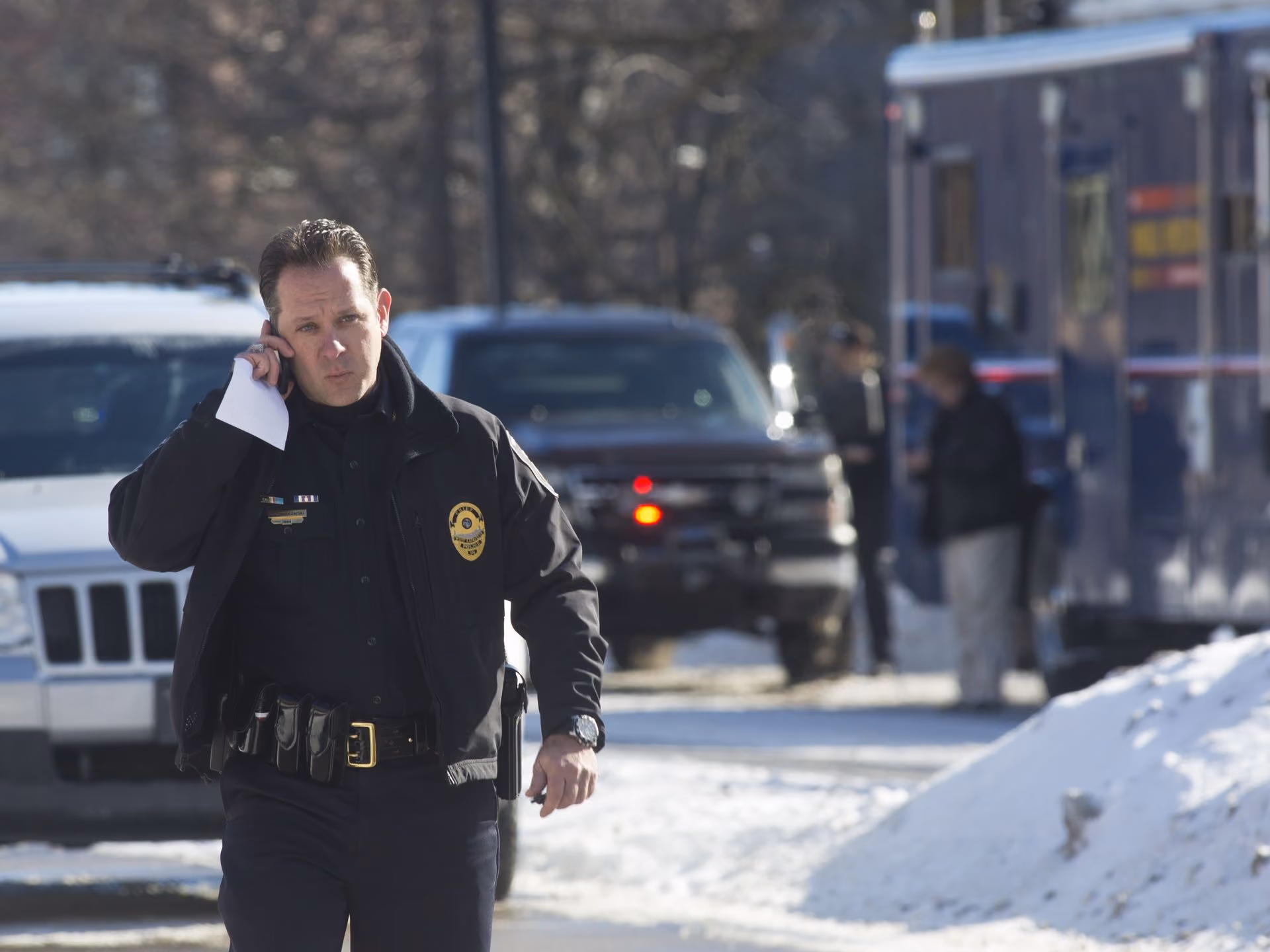 Police officer in uniform talking on phone outside in snowy area with vehicles and people in background.