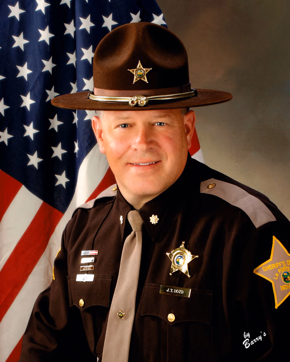 Smiling sheriff in uniform and hat posing in front of an American flag backdrop.