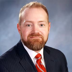 Man with red beard wearing a dark pinstripe suit and red striped tie, smiling against a gray background.