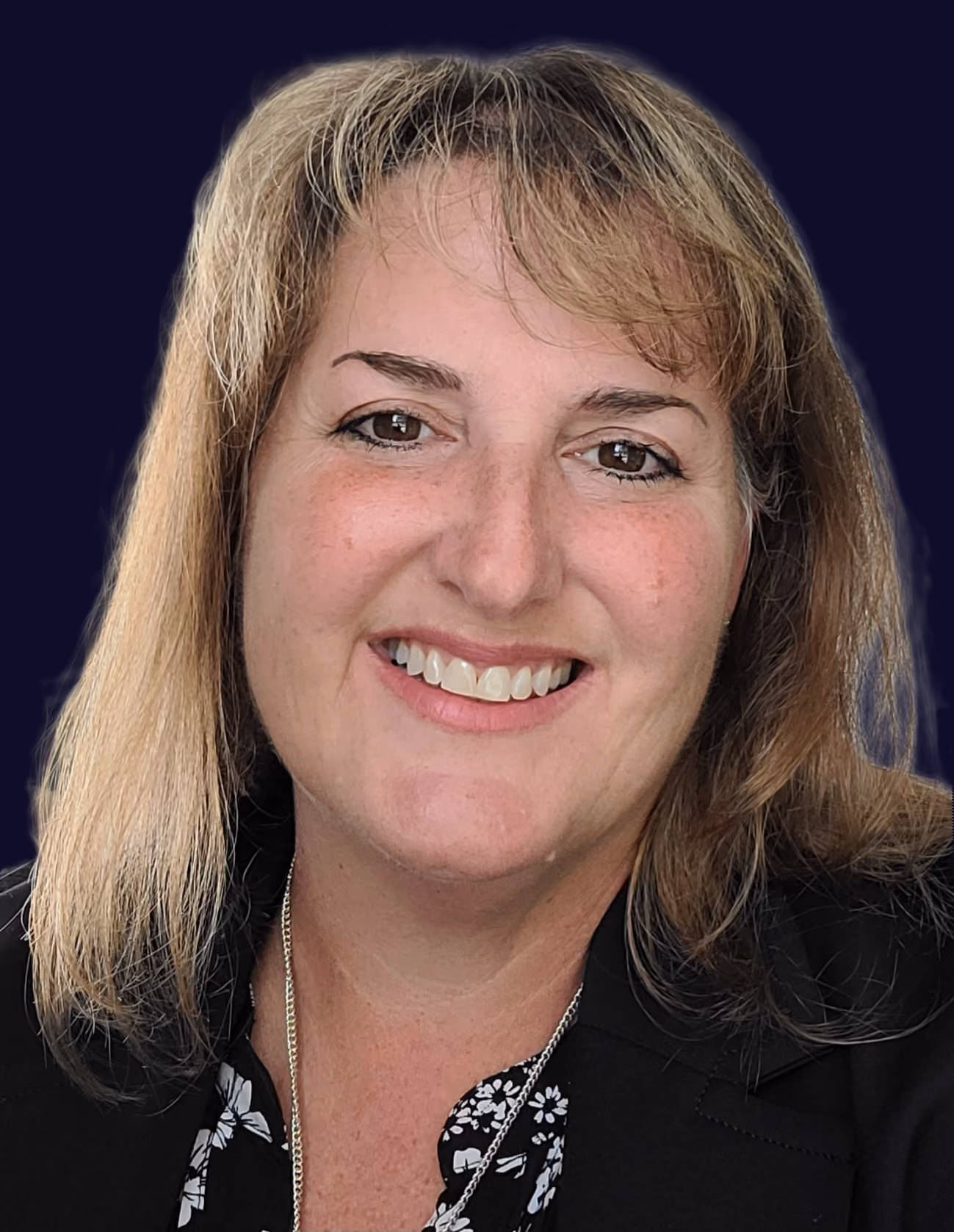 Smiling woman with shoulder-length blond hair wearing a black blazer and floral top on dark background.