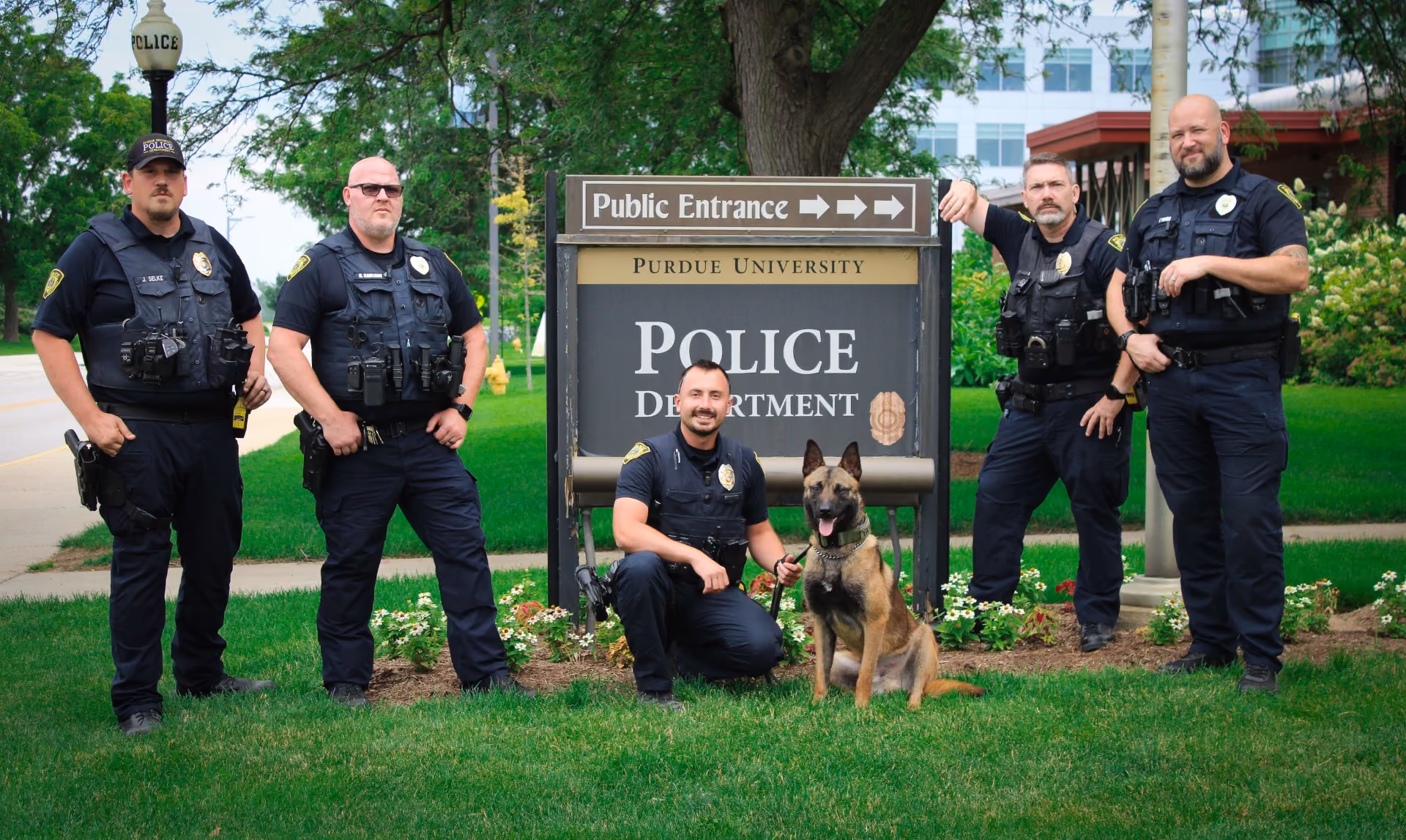 Five police officers in uniform with a K9 dog pose outside Purdue University Police Department sign on grass.