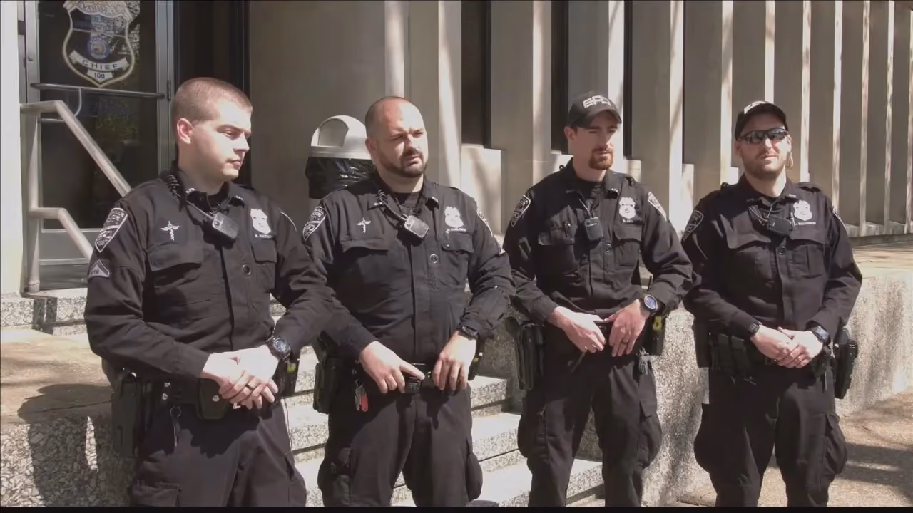 Four police officers in black uniforms standing in front of a building with steps and a badge emblem on the door.