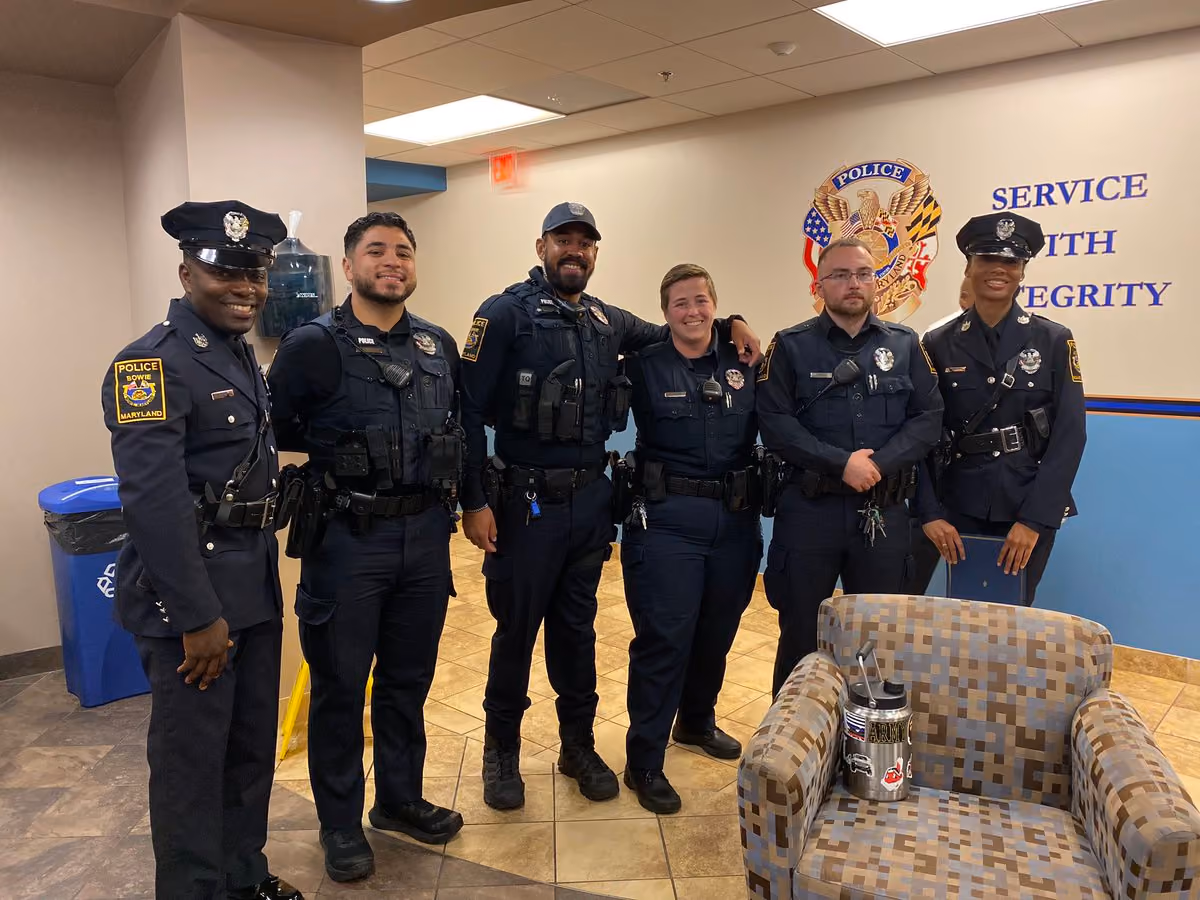 Six smiling police officers standing indoors in uniform in front of a wall with a police emblem and motto.