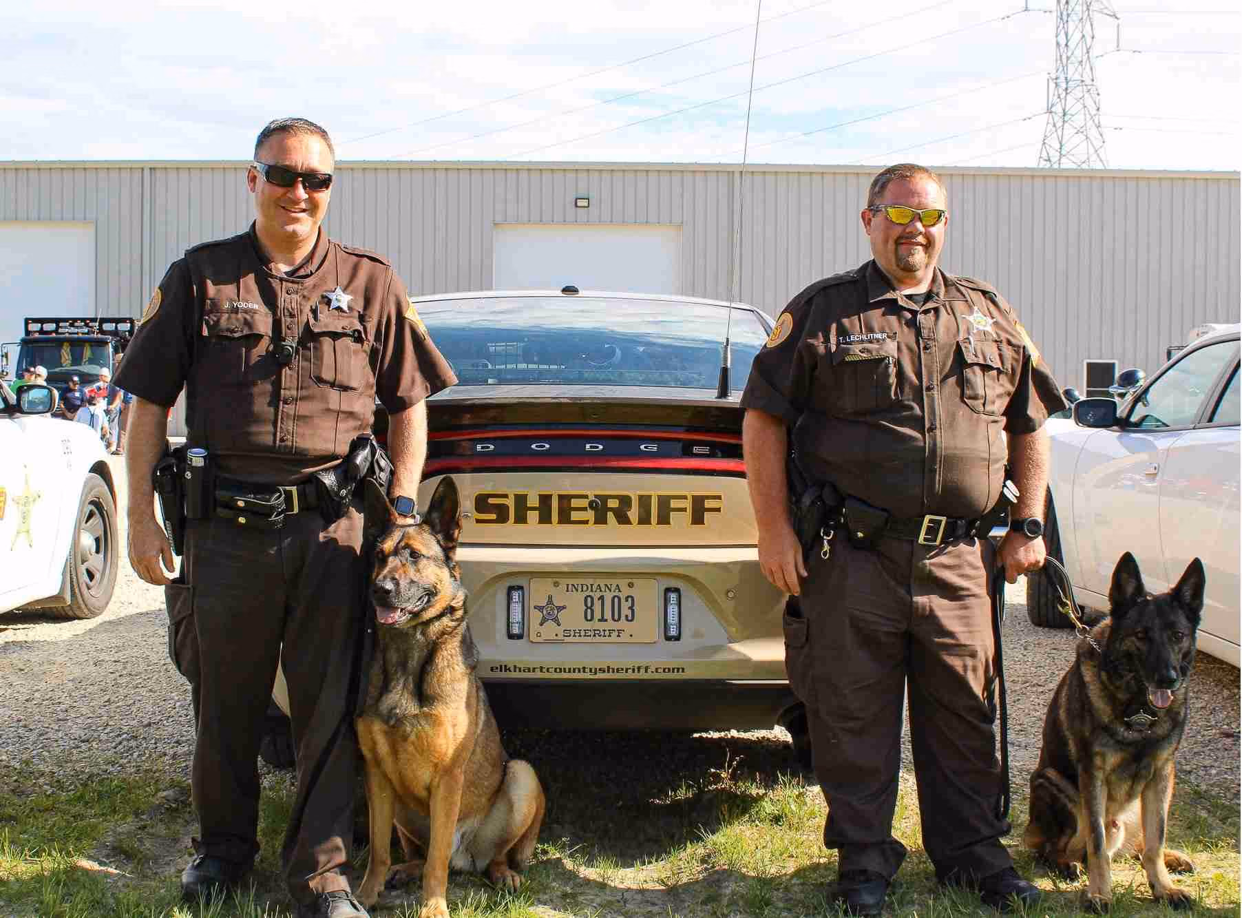 Two sheriff officers stand beside a sheriff car with their police German shepherd dogs on leashes.