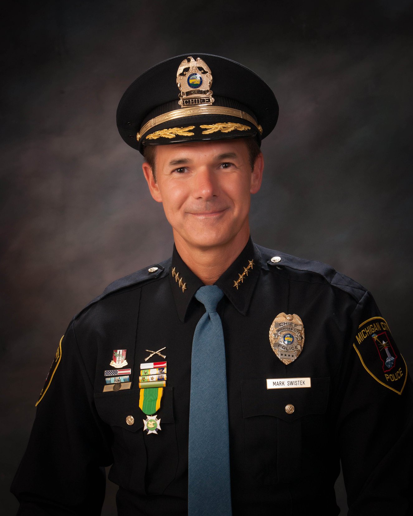 Police chief in uniform with badges, medals, and Michigan City Police patch on dark background.