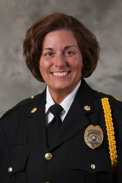 Smiling female police deputy chief in uniform with badge and gold cord on shoulder against gray background.