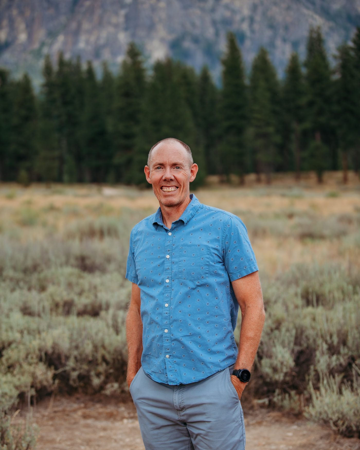 Greg Burnham standing outdoors with mountains and pines behind him