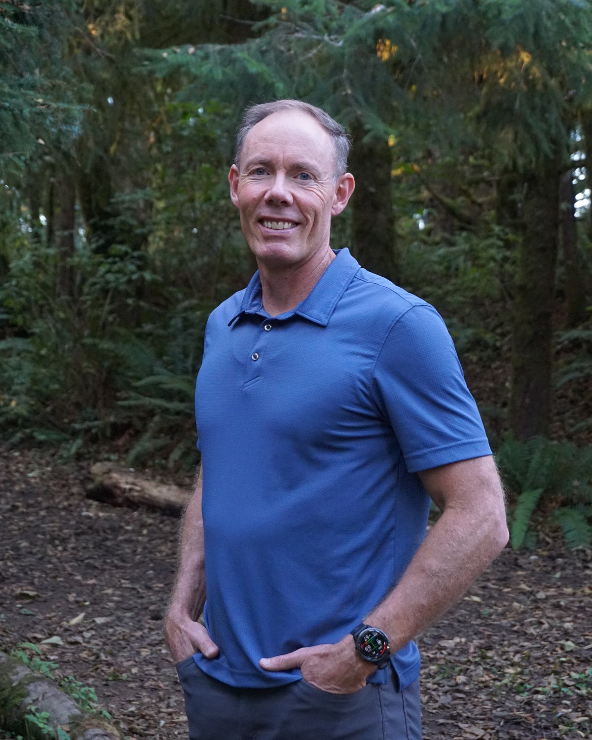 Greg Burnham smiling with his hands in his pockets in an Oregon forest.