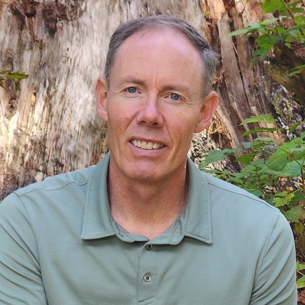Greg Burnham smiling in front of an old tree stump in an Oregon forest.