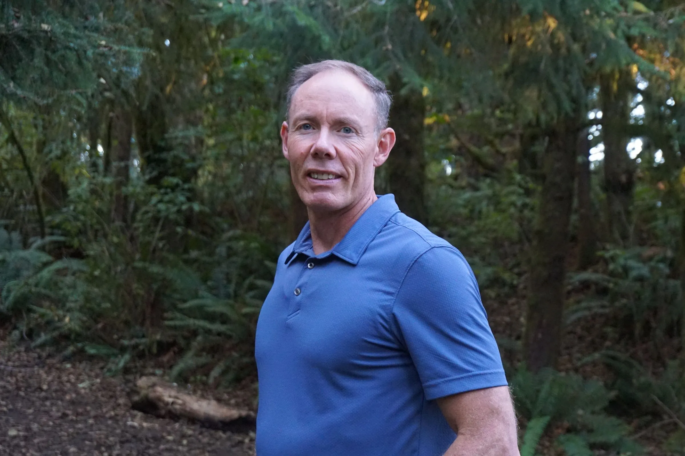 Greg Burnham smiling on a forest path among tall evergreen trees in Oregon.