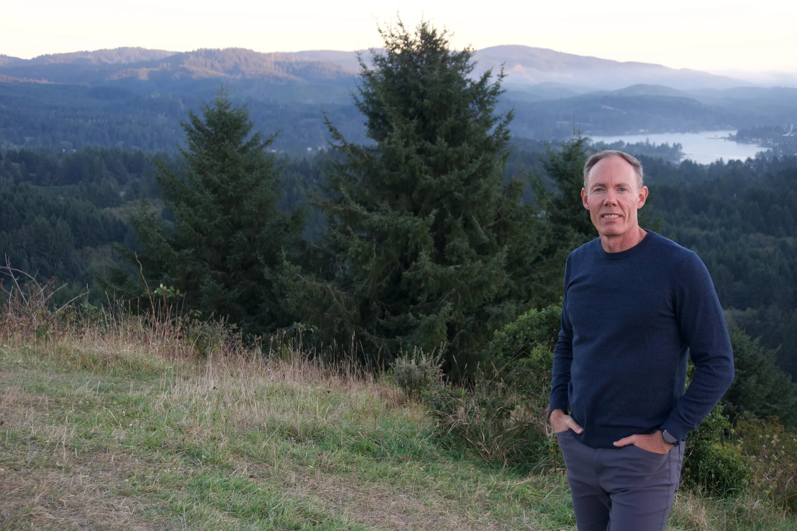 Greg Burnham standing on a grassy hillside above forested hills and a lake in Oregon.