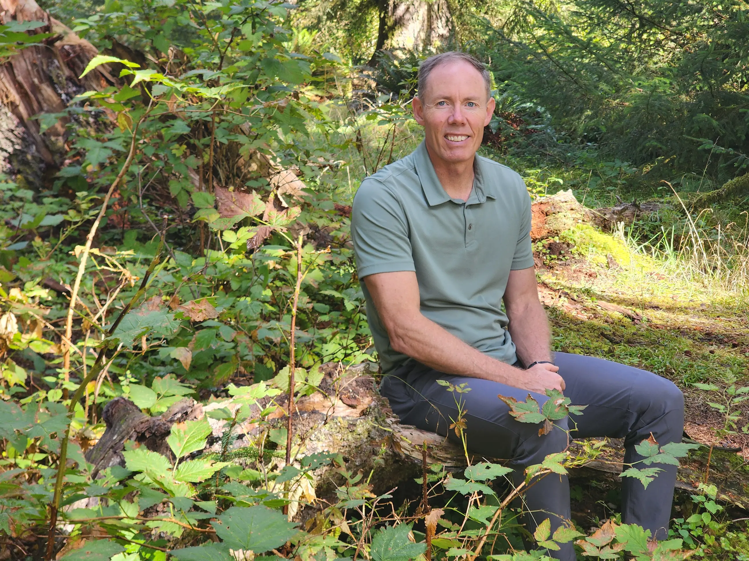 Greg Burnham smiling, seated on a log in a sunlit forest in Oregon.