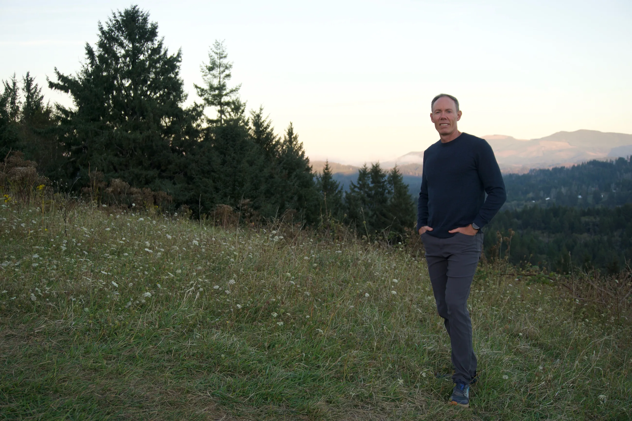 Greg Burnham standing in a grassy meadow at dusk, with evergreen trees and Oregon hills behind him.