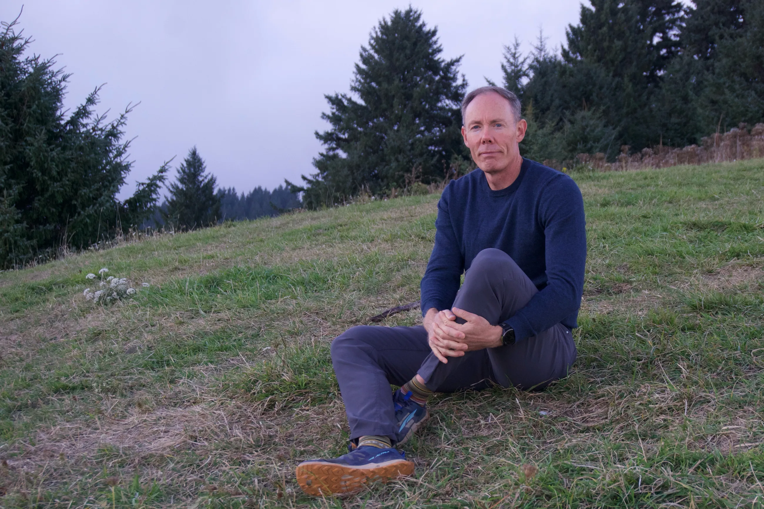 Greg Burnham sitting on a grassy hillside in Oregon, with evergreen trees behind him.