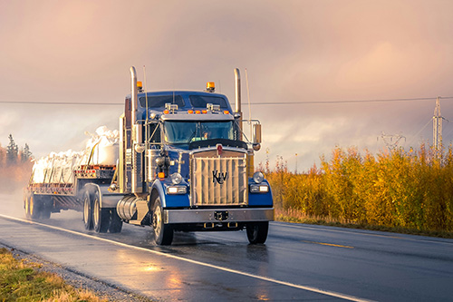 A blue semi-truck hauling a flatbed trailer drives down a wet highway surrounded by autumn trees under a golden evening sky.