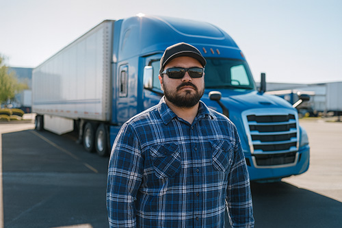 A truck driver wearing sunglasses and a blue plaid shirt stands confidently in front of a blue semi-truck parked in a lot on a sunny day.