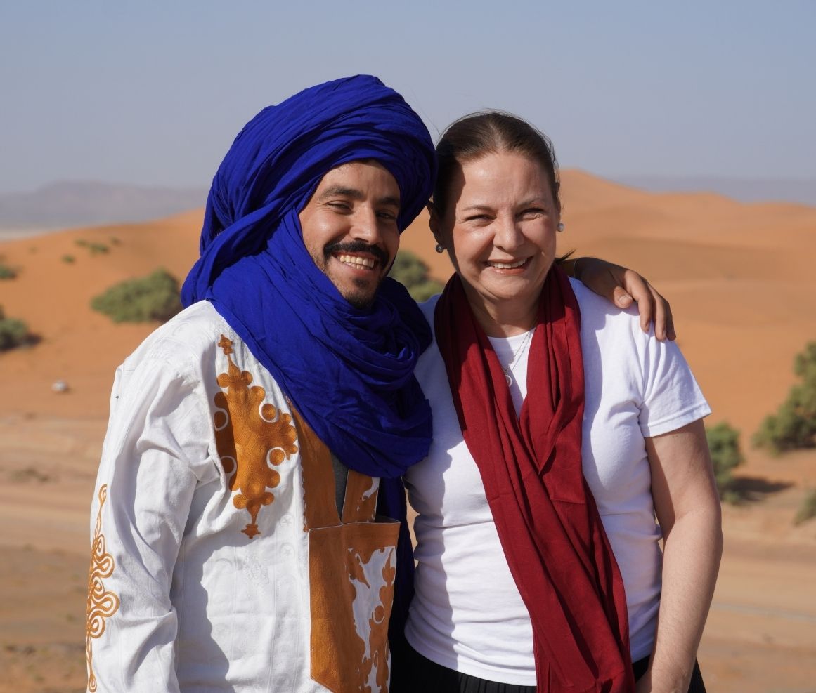 Hombre con turbante azul y ropa tradicional posa abrazado con una mujer que viste camiseta blanca y bufanda roja en un paisaje desértico.