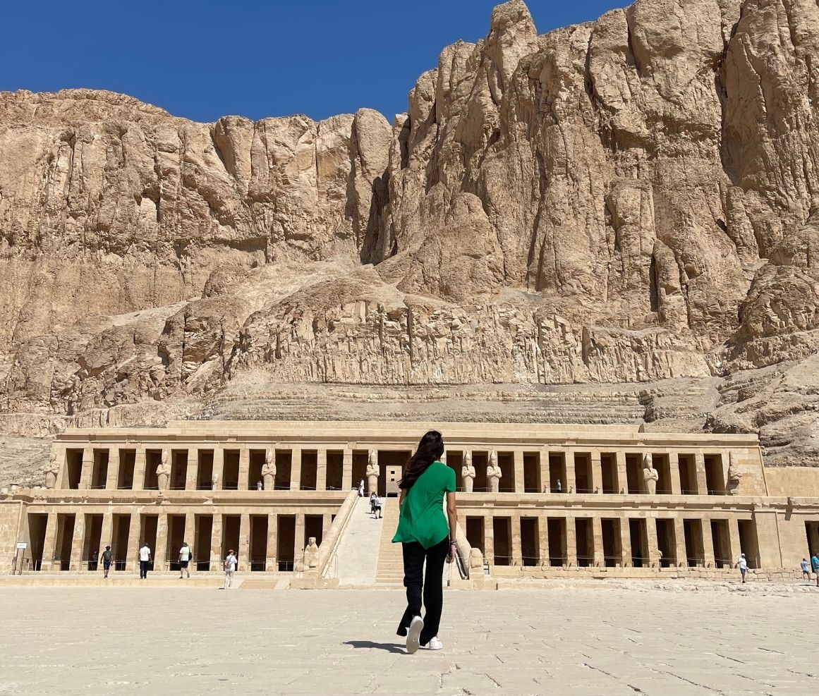 Persona de espaldas caminando hacia el templo de Hatshepsut ubicado frente a un acantilado rocoso bajo un cielo despejado azul.