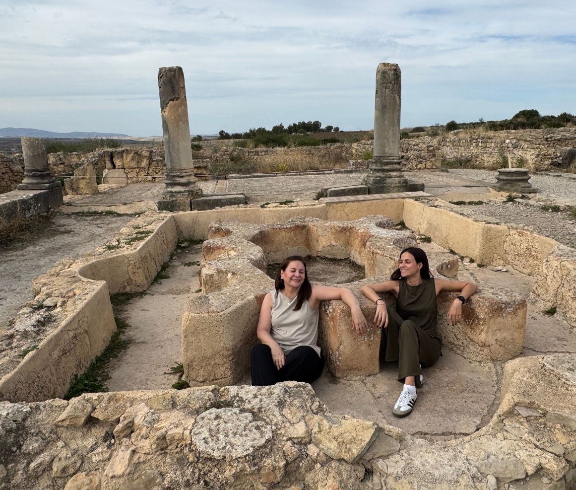 Dos mujeres sentadas y sonriendo dentro de ruinas arqueológicas con columnas antiguas y un cielo nublado.
