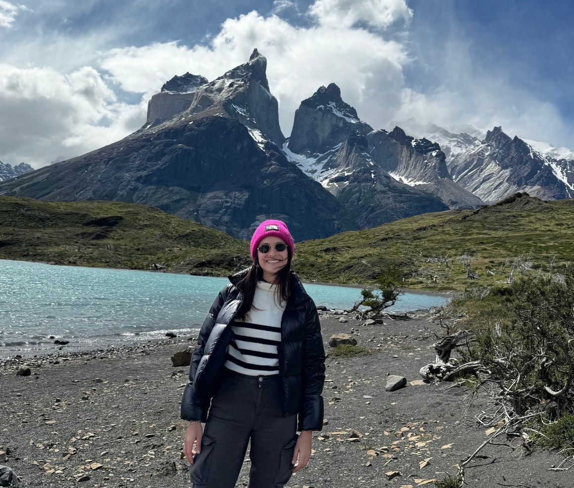 Mujer sonriente con gafas oscuras, gorro rosa y chaqueta negra de plumón frente a un lago azul y montañas rocosas con nieve bajo un cielo parcialmente nublado.