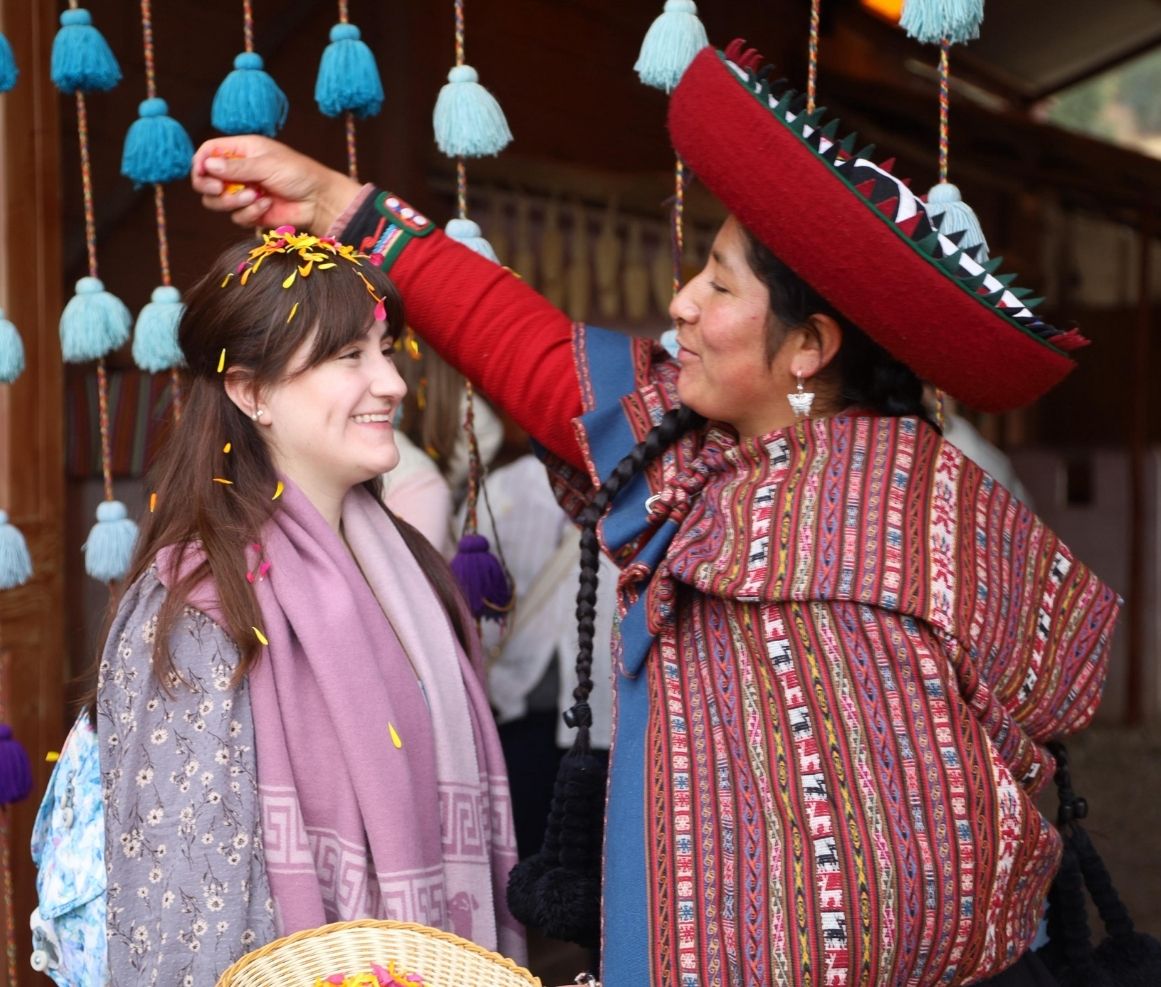 Mujer con vestido tradicional peruano esparce pétalos de flores sobre la cabeza de una joven sonriente que sostiene una canasta.