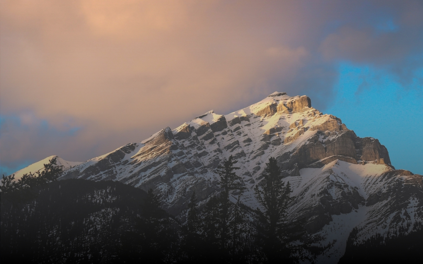Montaña nevada iluminada por la luz dorada del atardecer con árboles oscuros en primer plano y cielo parcialmente nublado.