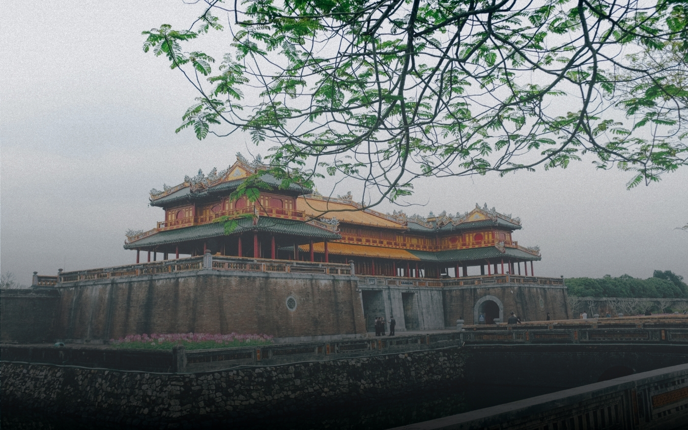Edificio histórico tradicional asiático con techo decorativo rojo y amarillo, rodeado por un muro de piedra y un puente, con ramas de árbol verdes en primer plano.