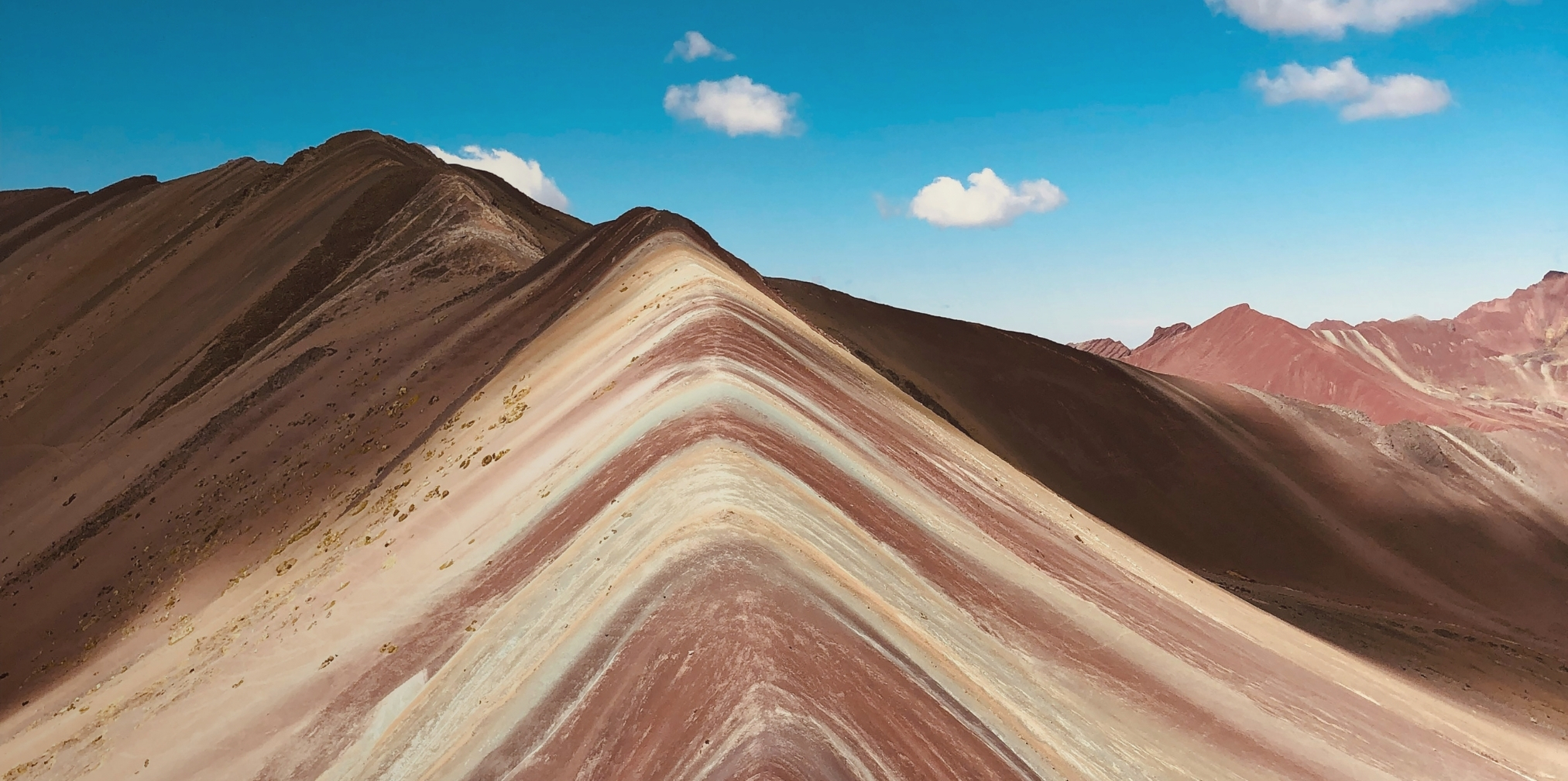 Montaña colorida con franjas naturales en tonos rojo, beige y marrón bajo un cielo azul con algunas nubes.