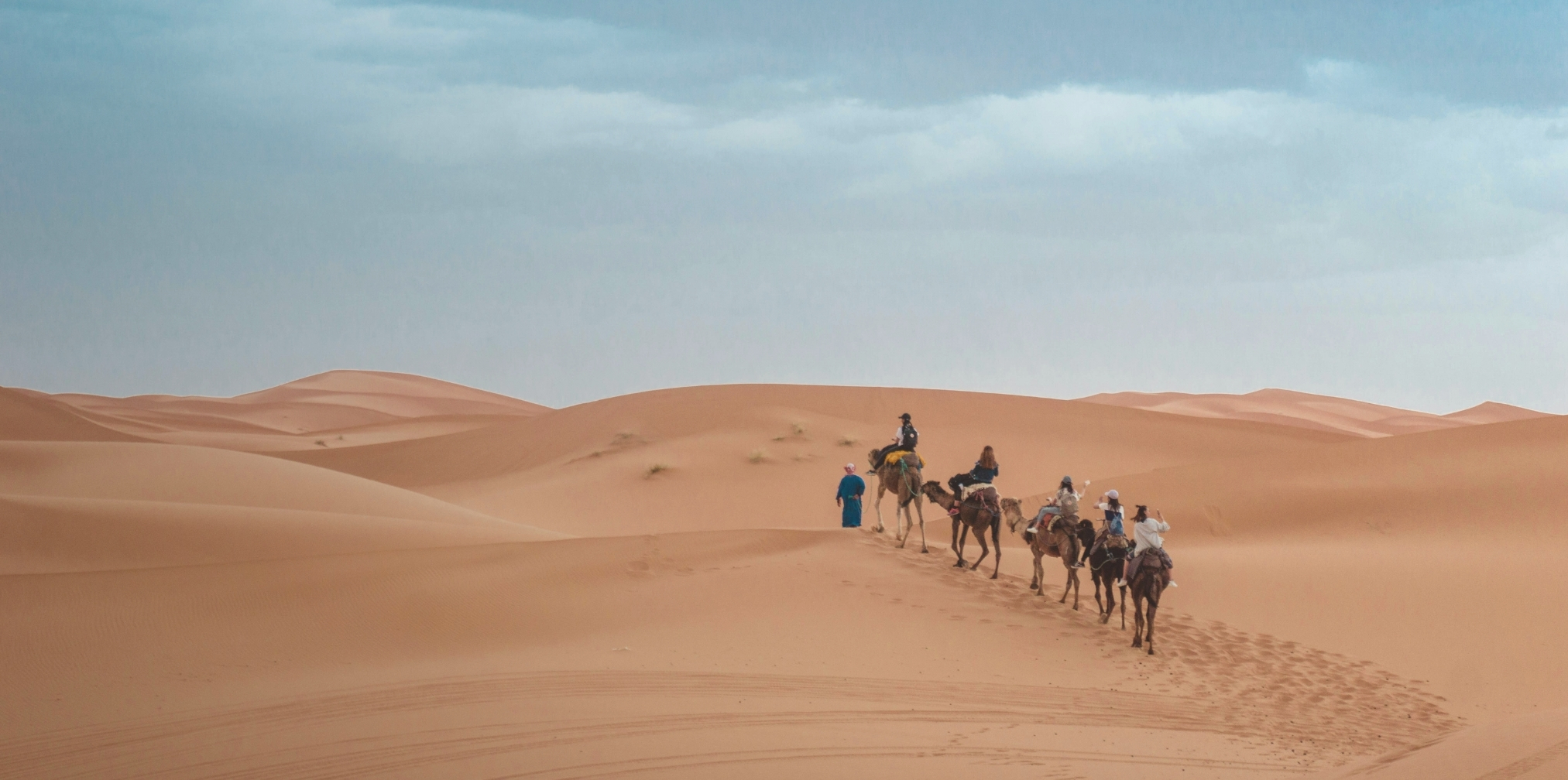 Caravana de turistas montando camellos que cruzan dunas de arena en un desierto bajo cielo nublado.