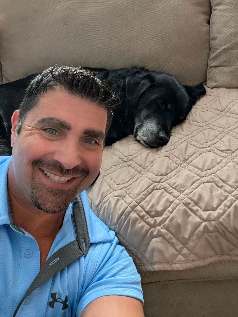 A man with a blue shirt and a black dog laying on a couch.