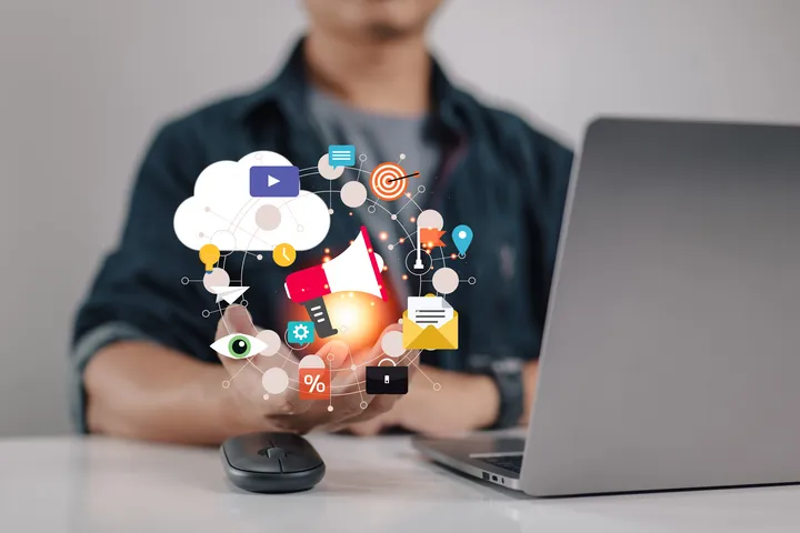 A man is sitting in front of a laptop with icons and a mouse.