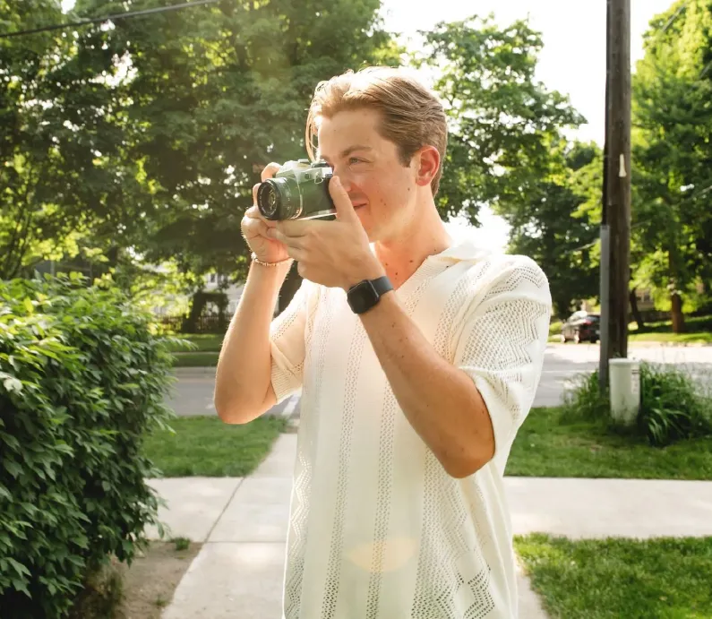 Young man in white shirt outdoors taking a photo with a camera on a sunny day.
