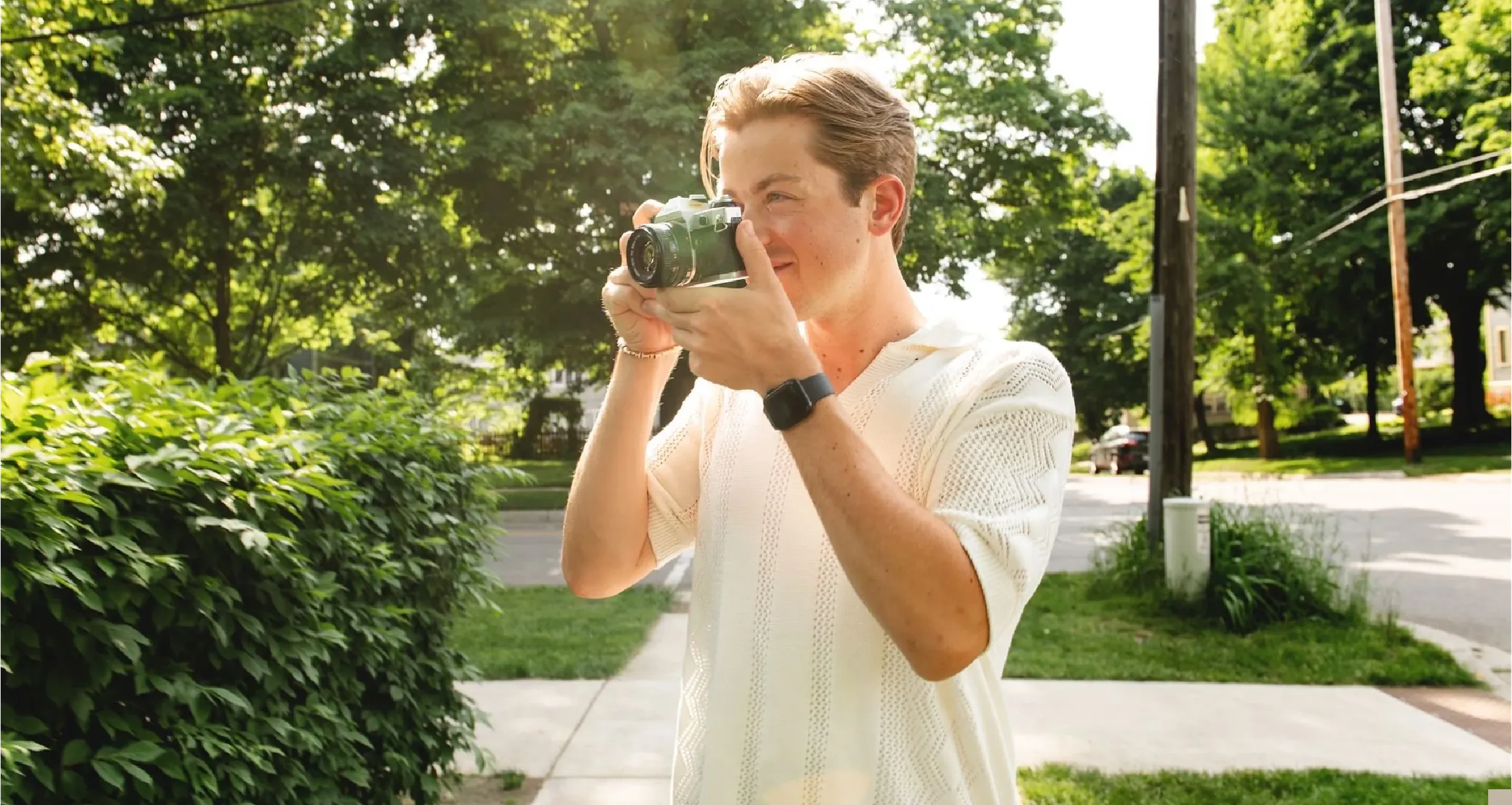 Man in a white short-sleeve shirt holding a camera and taking a photo outdoors on a sunny day.