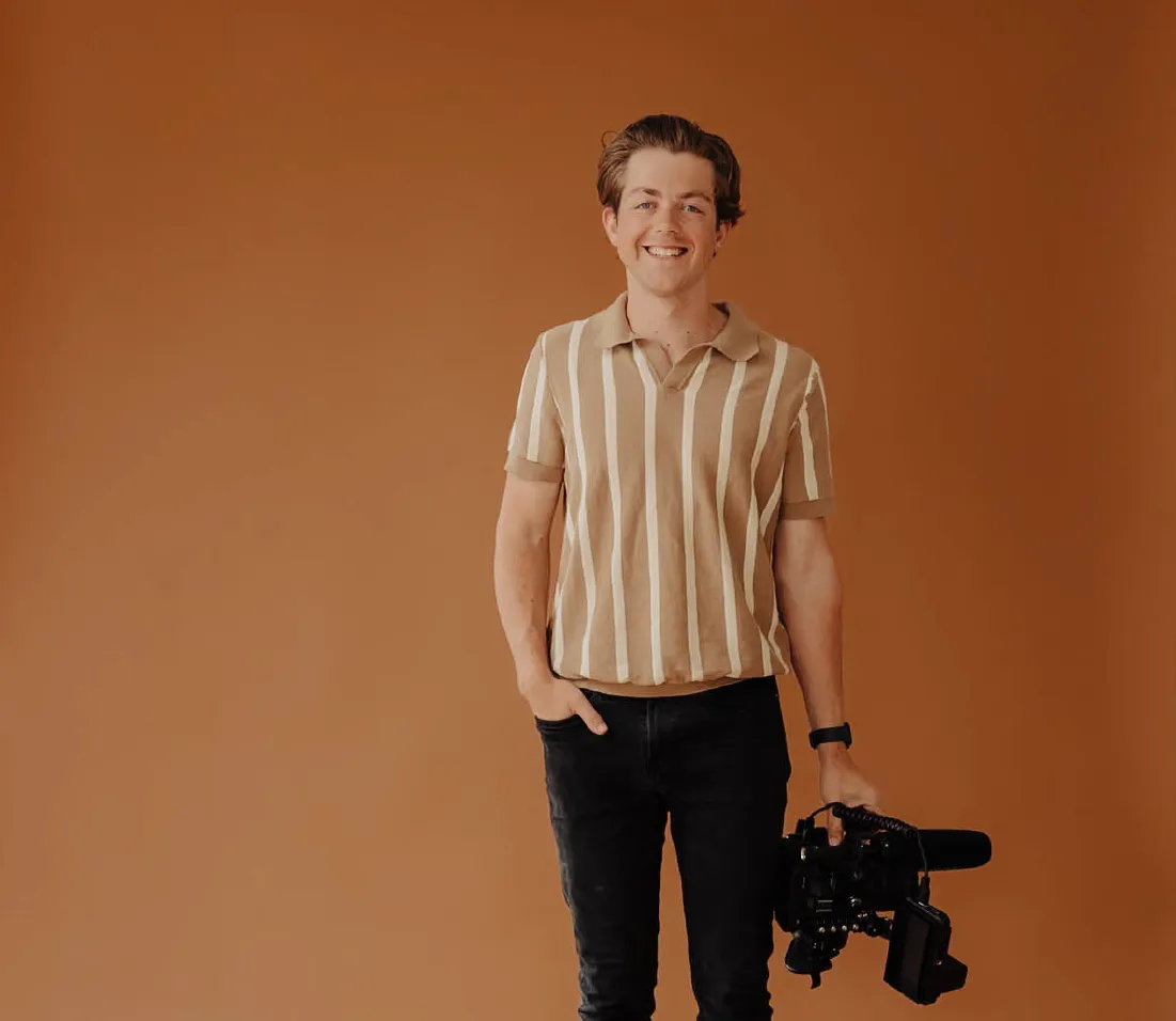 Smiling young man wearing a beige and white striped polo shirt holding a professional video camera against a brown background.