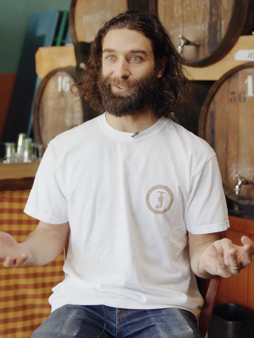 Bearded man with long curly hair in a white t-shirt talking animatedly in a brewery setting with wooden barrels in the background.