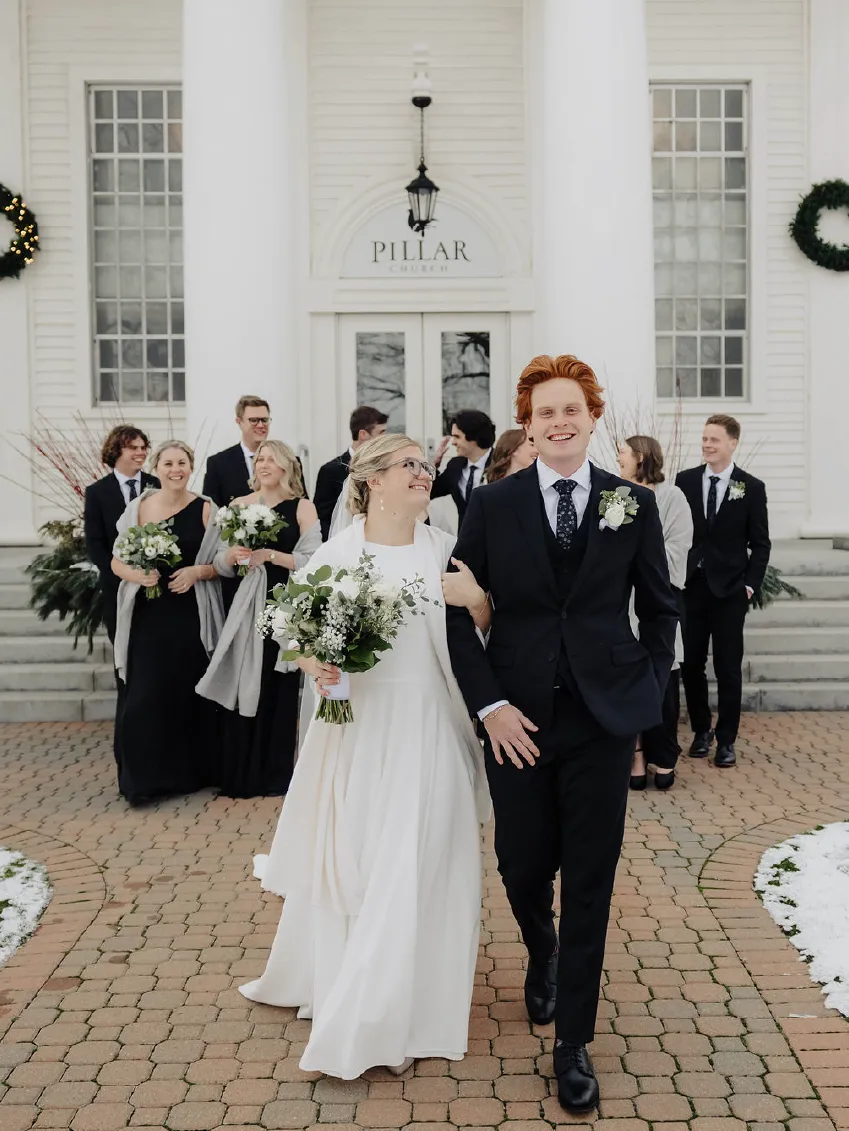 Bride and groom smiling and walking arm in arm outside Pillar Church with wedding party behind them.