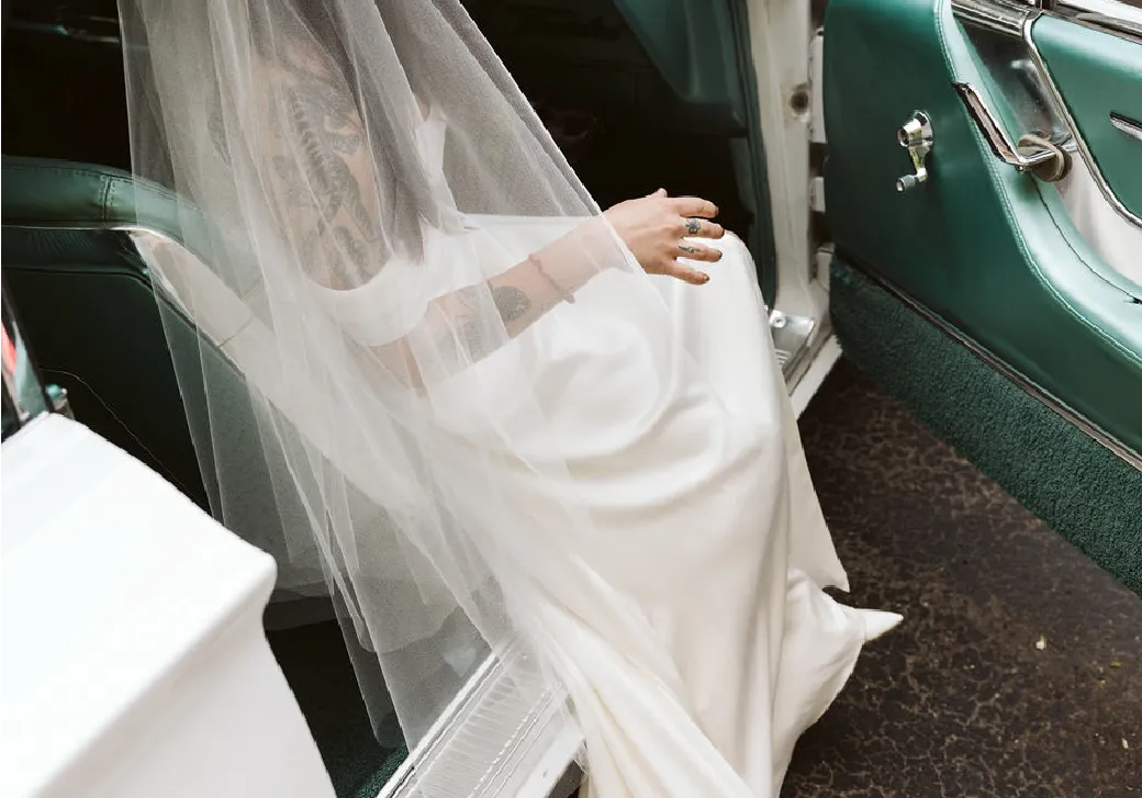 Bride with tattoos wearing a white wedding dress and veil seated inside a vintage car with green interior.