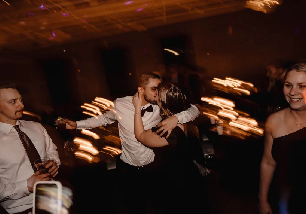 Couple kissing and embracing at a dimly lit party with other guests nearby.